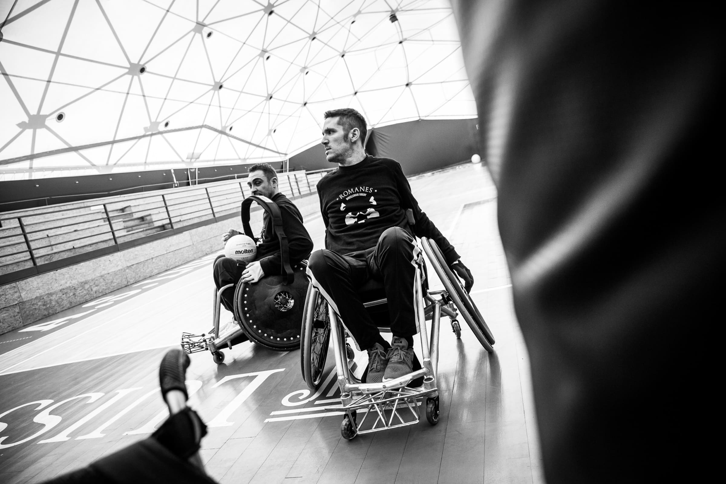 Due giocatori dei Romanes in carrozzina da rugby, fermi sul campo sotto la grande cupola prima dell'allenamento. Foto in bianco e nero.