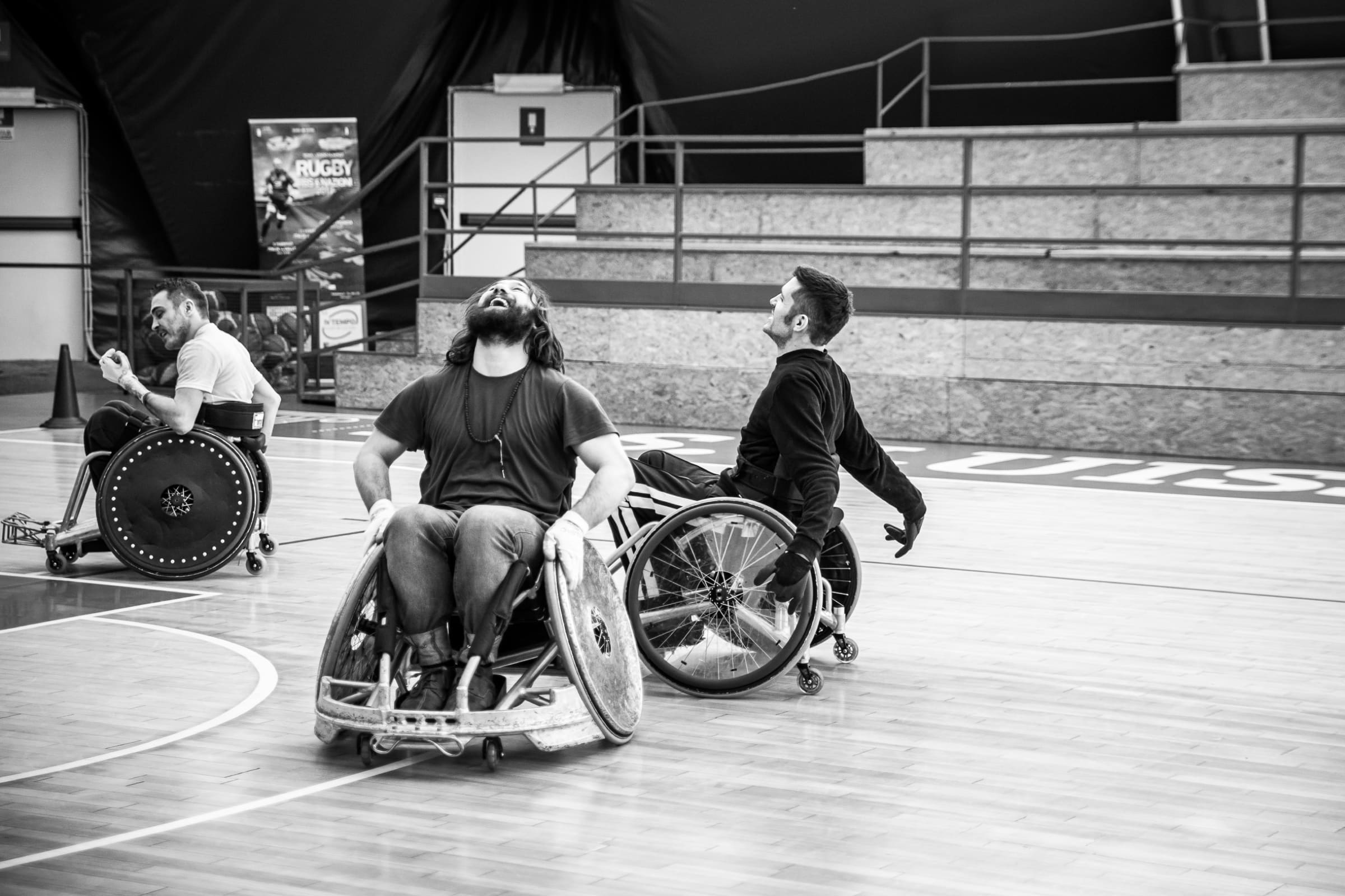 Tre giocatori dei Romanes ridono a testa alta durante un allenamento di wheelchair rugby. Foto in bianco e nero.